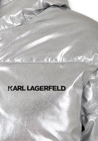 Giacca a vento argentata e lucida con texture trapuntata e logo nero "KARL LAGERFELD". Presenta una superficie liscia e riflettente.