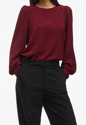 Frau trägt eine strukturierte, burgunderrote Langarmbluse mit gerafften Bündchen und hoch taillierte schwarze Hose, Hände in den Taschen.