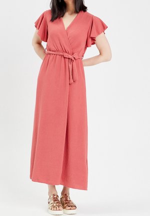 Kobieta w koralowej sukience kopertowej z falbanami na rękawach i paskiem przewiązanym w talii, w połączeniu z metalicznymi złotymi sandałami na platformie.