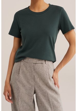 Kobieta ubrana w ciemnozielony t-shirt z krótkim rękawem i okrągłym dekoltem, wsunięty w beżowe, teksturowane spodnie z wysokim stanem i bocznymi kieszeniami.
