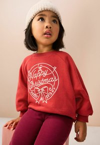 Bambino che indossa un maglione rosso con la scritta "Buon Natale Allegro e Splendente", cappello di lana e pantaloni viola, seduto che guarda leggermente verso l'alto.