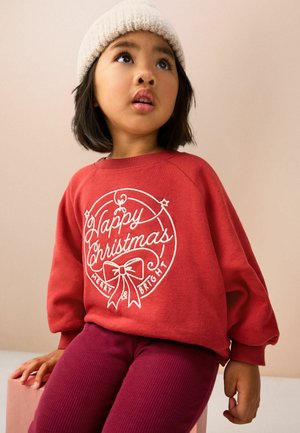 Ein kleines Kind trägt einen roten Pullover mit der Aufschrift "Frohe Weihnachten, fröhlich und hell", eine gestrickte Mütze und lila Hosen. Es sitzt und schaut leicht nach oben.