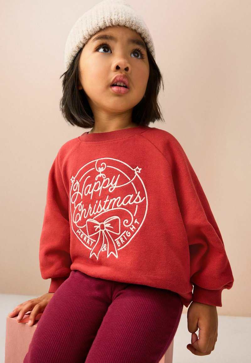 Bambino che indossa un maglione rosso con la scritta "Buon Natale Allegro e Splendente", cappello di lana e pantaloni viola, seduto che guarda leggermente verso l'alto.