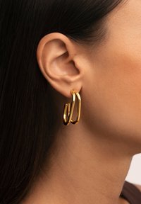 Orecchini a cerchio d'oro esposti sull'orecchio, caratterizzati da un design elegante e lucido con forme intrecciate e una superficie liscia.