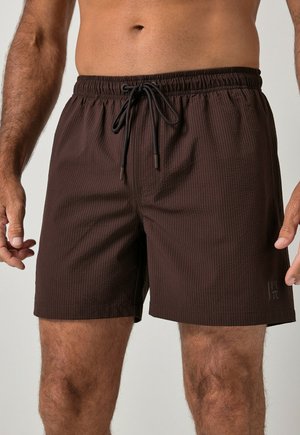 Man die donkerbruine gestructureerde short draagt met elastische tailleband en zwart trekkoord, getoond van taille tot midden dij.