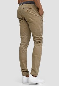 Chinos caqui de algodón con corte recto, que cuentan con bolsillos laterales, un bolsillo trasero con cremallera y una cinturilla estampada, combinados con zapatillas blancas.