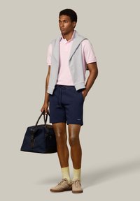 Mężczyzna w różowej koszulce polo, granatowych szortach, żółtych skarpetkach, beżowych trampkach, szarej bluzie przewieszonej przez ramiona, trzymający granatową torbę sportową.