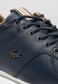 Zapatilla de cuero azul marino con cordones azul oscuro, ojales metálicos, etiqueta de lengua a rayas en beige y blanco, y logo de cocodrilo plateado en el lateral.