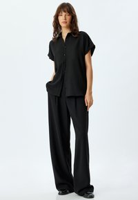 Camisa negra de manga corta y botones combinada con pantalones de pierna ancha de tiro alto negros y zapatos negros, que presentan una tela texturizada.