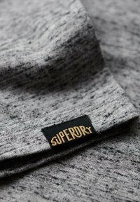 Gray fabric with a textured pattern, featuring a black label that reads "SUPERDRY" in gold letters, positioned along the seam.