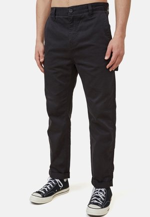 Mand iført sorte bukser med manchetter og sorte høje sneakers med hvide snørebånd, stående mod en ensfarvet baggrund.