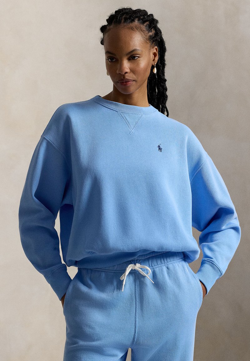 Ανοιχτό μπλε φούτερ με στρογγυλή λαιμόκοψη και χαμηλωμένους ώμους, με μικρό λογότυπο στο στήθος. Μαλακό ύφασμα με ριμπ μανσέτες.