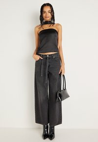 Top preto de cetim sem alças, combinado com jeans de ganga preta de cintura alta e perna larga. A modelo segura uma pequena bolsa preta; botas de salto pretas brilhantes.