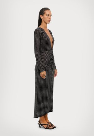 Black, long-sleeve dress with a deep V-neck, featuring a polka dot pattern, cinched waist, and ankle-length hem. Paired with strappy black heels.