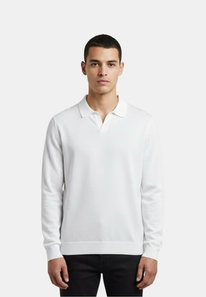 Giovane uomo che indossa una polo bianca semplice a maniche lunghe e pantaloni neri, in piedi davanti a uno sfondo chiaro e neutro.