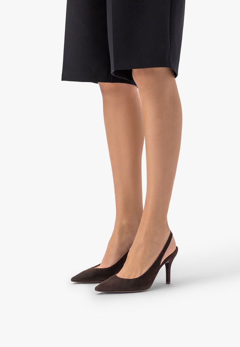Jambes portant un short noir jusqu'aux genoux et des escarpins slingback en daim marron à bout pointu avec des talons aiguilles de hauteur moyenne sur fond blanc.