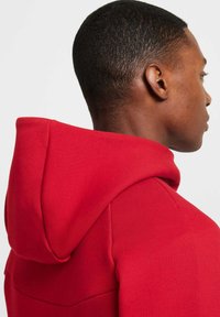 Sudadera roja con capucha de textura suave, que presenta un escote en ángulo y una capucha prominente. La tela parece suave y resistente.