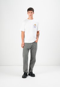 Weißes T-Shirt mit kurzen Ärmeln und buntem Grafikdesign, kombiniert mit grauen geraden Jeans und schwarzen Schuhen mit dickem Sohlen.