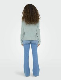 Camisa de manga larga verde claro combinada con jeans acampanados azules, mostrando la textura y el ajuste desde la parte de atrás, con cabello rizado visible.