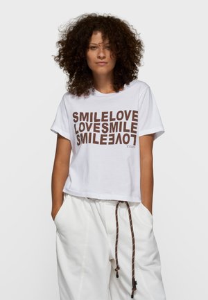 Biały krótki t-shirt z bawełny z brązowym napisem "SMILELOVE LOVESMILE" w pogrubionym, powtarzającym się wzorze. Krótkie rękawy, luźny krój.