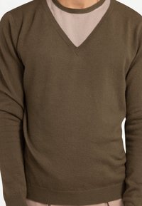 Brun V-ringad tröja i ett mjukt stickat tyg, med långa ärmar och texturerad yta, lager-på-lager över en ljusfärgad undertröja.