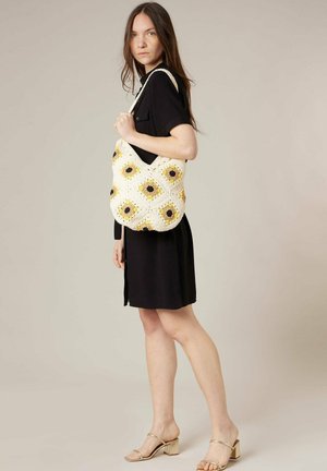 Femme en robe noire tenant un sac d'épaule en crochet crème avec des motifs de tournesols jaunes et marron, portant des sandales à talons blocs beige.