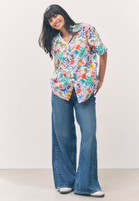 Kurzärmliges, mehrfarbiges Hemd mit Blumenmuster, kombiniert mit weitem blauen Denim-Jeans und weißen Turnschuhen.