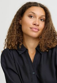 Camicia nera con collare, dalla texture liscia, dotata di chiusura a bottoni e una leggera lucentezza, indossata da una persona con capelli ricci.