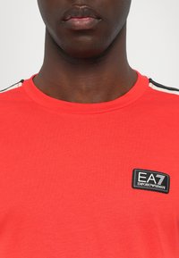 Red cotton t-shirt featuring a black EA7 logo patch on the chest and contrasting black and white stripes on the shoulder. Smooth texture.