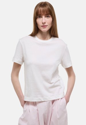 Jeune femme aux cheveux roux lisses portant un t-shirt blanc uni à manches courtes et un pantalon rose clair, mains dans les poches, fond neutre.