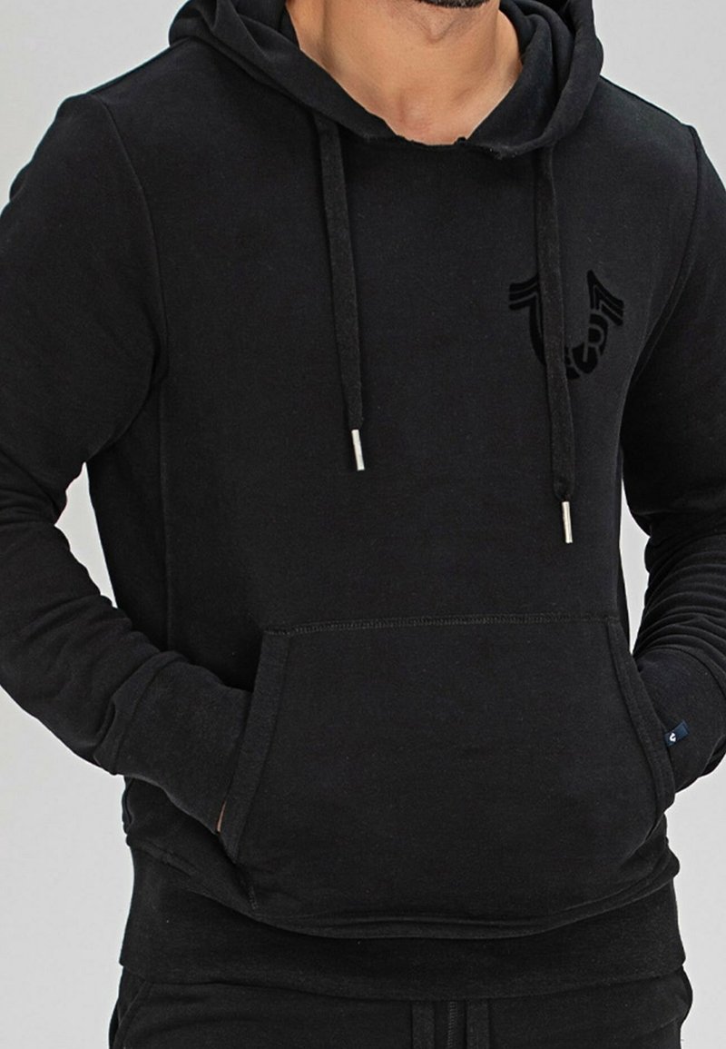 Homme portant un sweat à capuche noir avec poche frontale et cordons, mains dans les poches, vue partielle du torse et du cou.