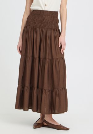 Donna che indossa una lunga gonna marrone a balze con una fascia elastica arricciata in vita, abbinata a ballerine marroni con cinturino sul tallone, in piedi davanti a uno sfondo semplice.