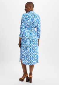 Blauwe jurk met patronen, lange mouwen, een cinched taille en zij-splitten. Het ontwerp bevat ingewikkelde witte motieven op een blauwe achtergrond.