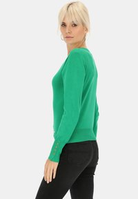 Zielony sweter z długim rękawem i dekoltem w kształcie litery V, ze ściągaczem na dole oraz trzema guziczkami przy mankietach. Miękka tekstura i dopasowany krój. Na sobie czarne dżinsy.