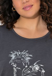 Dunkelgraues T-Shirt mit einer weißen grafischen Illustration von zwei Vögeln mit markanten Augen und detaillierten Federn, runder Ausschnitt und weichem Stoff.