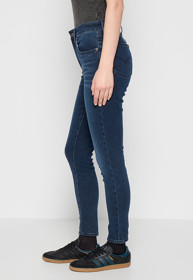 Schmale dunkelblaue Jeans mit hoher Taille, glatter Textur und zwei Gesäßtaschen. Kombiniert mit schwarzen Sportschuhen mit blauen Akzenten.