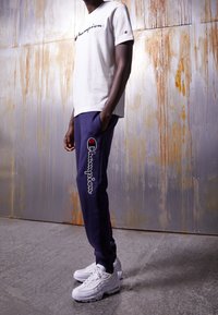 Bílé tričko s krátkým rukávem s logem "Champion", doplněné tmavě modrými tepláky s vyšívaným logem. Model má na sobě bílé tenisky.