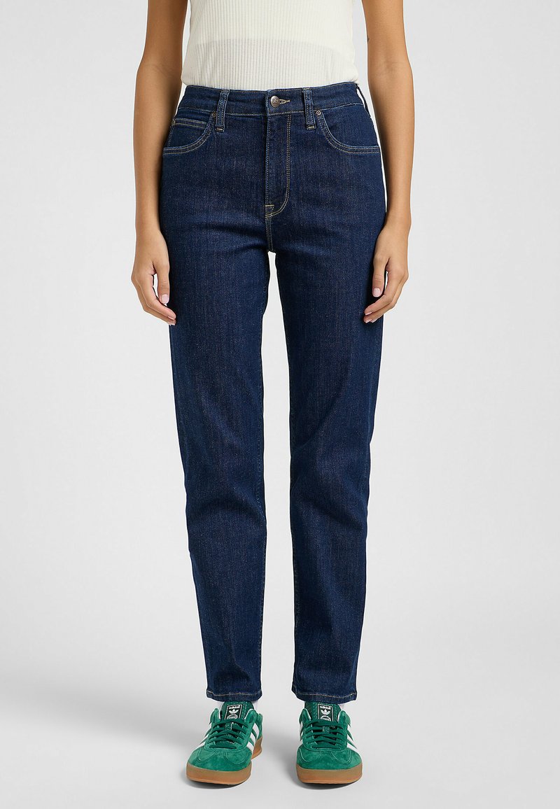 Pantalones vaqueros de mezclilla azul oscuro con un diseño de pierna recta, que cuentan con cinco bolsillos, costuras claras y una cintura alta, combinados con zapatillas verdes.