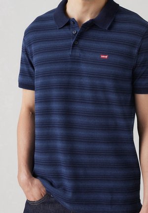 Homme portant un polo bleu marine à rayures à manches courtes avec col, deux boutons et logo rouge Levi's sur la poitrine, main dans la poche du jean.