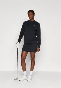 Woman standing with golf club wearing black long-sleeve top, black skirt, white golf glove, white socks, and white sneakers on plain background.