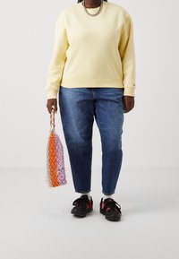 Sweatshirt jaune avec logo blanc, jeans droits bleus et chaussures noires avec accents rouges, tenant un sac en filet multicolore.