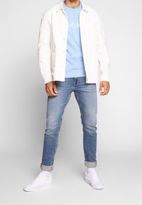 Homme portant un t-shirt bleu clair de Calvin Klein, une veste blanche boutonnée, un jean bleu retroussé, des chaussettes blanches et des baskets blanches, debout.