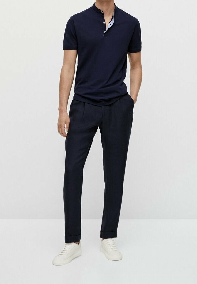 Homme portant un t-shirt henley bleu marine à manches courtes, un pantalon bleu marine foncé et des baskets blanches, debout avec une main dans la poche.