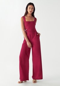 Ženska v brezrokavnem, širokem, temno roza kombinezonu z pasom in kvadratnim izrezom, stoji z eno roko v žepu in nosi bež sandale.