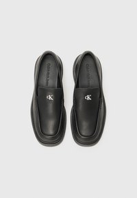 Scarpe slip-on nere in pelle liscia con punta arrotondata, caratterizzate da un design cucito e un piccolo logo "cK" argentato sul davanti.