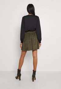 Haut noir à manches longues associé à une jupe olive longueur genou. La jupe possède une taille élastique et une texture douce semblable au daim. Bottes à talons noires.