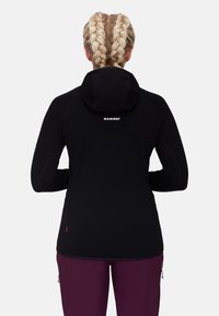 Black fleece jacket with a hood, featuring a logo printed on the back. Long sleeves with contrasting stitching, paired with deep purple pants.
