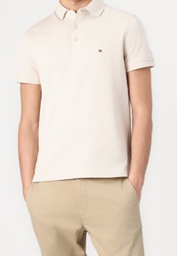 Polo beige en coton, avec un col et trois boutons, comportant un petit logo rouge et bleu sur la poitrine gauche.
