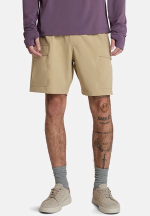 Pantalones cortos beige con dos bolsillos laterales tipo cargo, confeccionados en un material ligero. El modelo lleva una camisa de manga larga morada y calcetines gris claro.