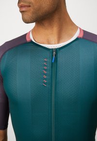 Maillot de cyclisme teal à manches courtes, doté d'une fermeture éclair à l'avant, d'un tissu texturé et d'accents roses et violets sur les épaules.